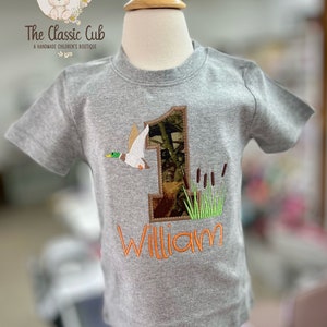 May include: A gray short-sleeved shirt with a camouflage number one and a duck applique. The name "William" is embroidered in orange below the number.