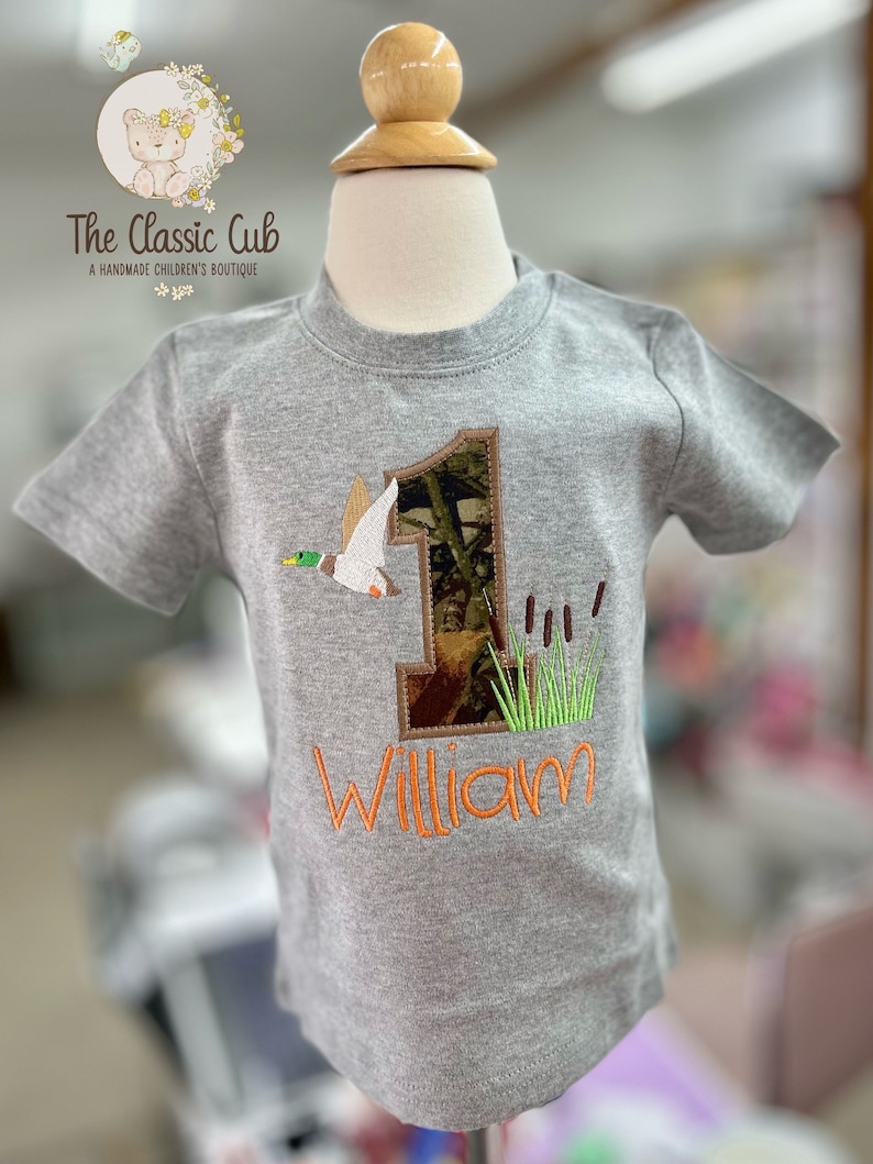 May include: A gray short-sleeved shirt with a camouflage number one and a duck applique. The name "William" is embroidered in orange below the number.