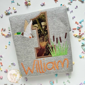 May include: A gray t-shirt with a camouflage number one, a duck flying, and green reeds embroidered on it. The name "William" is embroidered in orange below the number one.