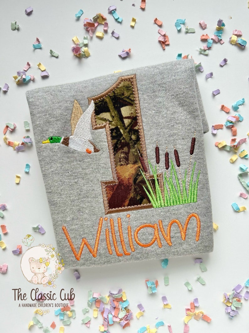 May include: A gray t-shirt with a camouflage number one, a duck flying, and green reeds embroidered on it. The name "William" is embroidered in orange below the number one.