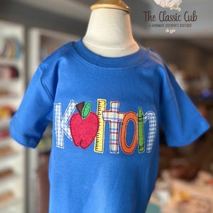 Puede incluir: Una camiseta azul de manga corta con el nombre "Kolton" aplicado en parches coloridos con tem&aacute;tica escolar. Los parches incluyen una manzana, una regla y un cray&oacute;n. La camiseta se muestra en un maniqu&iacute;.