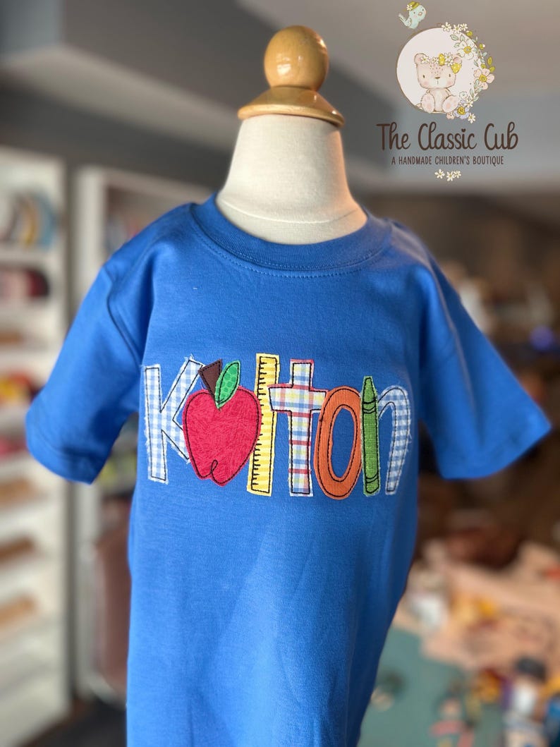 Puede incluir: Una camiseta azul de manga corta con el nombre "Kolton" aplicado en parches coloridos con tem&aacute;tica escolar. Los parches incluyen una manzana, una regla y un cray&oacute;n. La camiseta se muestra en un maniqu&iacute;.