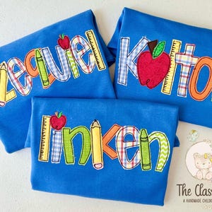 Puede incluir: Tres camisetas azules con nombres bordados, manzanas, l&aacute;pices y reglas. Los nombres son "Ezekiel", "Kitton" y "Linken". El logotipo de The Classic Cub est&aacute; en la esquina inferior derecha.
