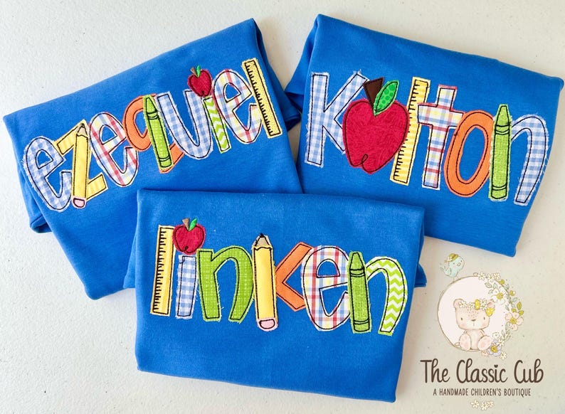 Puede incluir: Tres camisetas azules con nombres bordados, manzanas, l&aacute;pices y reglas. Los nombres son "Ezekiel", "Kitton" y "Linken". El logotipo de The Classic Cub est&aacute; en la esquina inferior derecha.