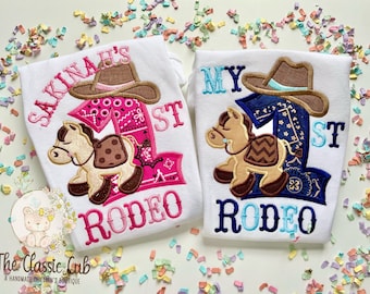 May include: Two white baby grows with embroidered designs. One grow says "Sakinah's 1st Rodeo" in pink with a pink bandana, brown horse, and brown cowboy hat. The other grow says "My 1st Rodeo" in blue with a blue bandana, brown horse, and brown cowboy hat.