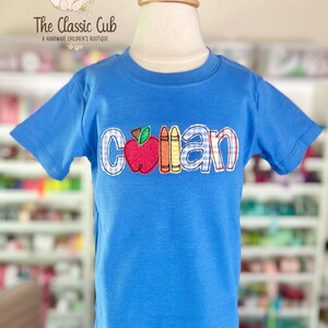 Puede incluir: Una camiseta azul de manga corta con el nombre "Collan" bordado en un patr&oacute;n de cuadros, con un dise&ntilde;o de manzana y l&aacute;pices de colores. La camiseta se muestra en un maniqu&iacute;. The Classic Cub, A Handmade Children's Boutique, est&aacute; impreso en la parte superior izquierda.