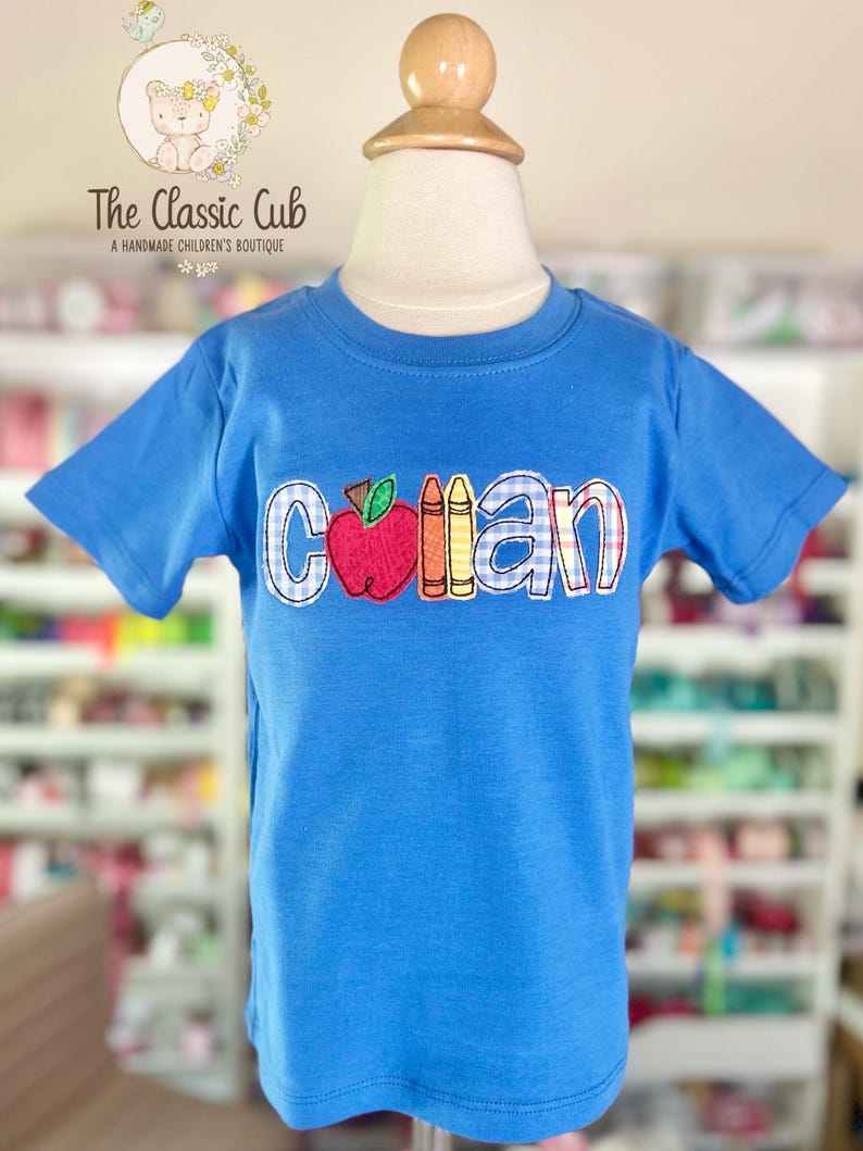 Puede incluir: Una camiseta azul de manga corta con el nombre "Collan" bordado en un patr&oacute;n de cuadros, con un dise&ntilde;o de manzana y l&aacute;pices de colores. La camiseta se muestra en un maniqu&iacute;. The Classic Cub, A Handmade Children's Boutique, est&aacute; impreso en la parte superior izquierda.