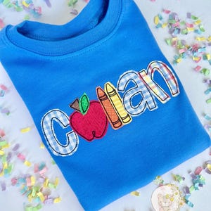 Puede incluir: Sudadera azul de cuello redondo con el nombre "Colan" aplicado en una mezcla de cuadros y colores lisos. Una manzana y l&aacute;pices de colores est&aacute;n aplicados junto al nombre. El logotipo de The Classic Cub es visible.