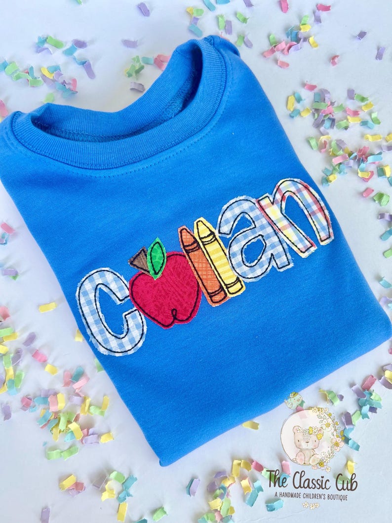 Puede incluir: Sudadera azul de cuello redondo con el nombre "Colan" aplicado en una mezcla de cuadros y colores lisos. Una manzana y l&aacute;pices de colores est&aacute;n aplicados junto al nombre. El logotipo de The Classic Cub es visible.