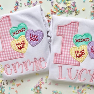 Peut inclure: Deux t-shirts blancs avec des chiffres un et un en tissu vichy rose et blanc, avec trois cœurs de conversation en bonbons qui disent "XOXO", "Hug Me" et "Luv You". Les t-shirts sont personnalisés avec les noms "Emmie" et "Lucy".