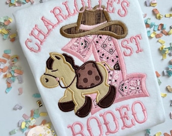 Puede incluir: Camiseta blanca con texto bordado en rosa y temática de rodeo. Presenta un sombrero de vaquero marrón, un pañuelo rosa y un caballo de dibujos animados. El texto dice "CHARLOTTE'S 1ST RODEO".