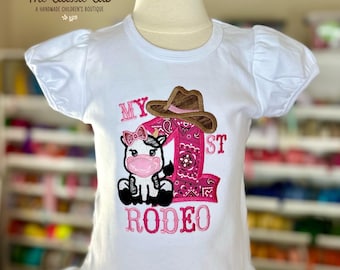 May include: White short-sleeved shirt with a pink and brown embroidered design featuring a cow wearing a cowboy hat and the text "My 1st Rodeo".