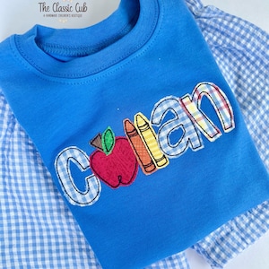 Puede incluir: Una camiseta azul para ni&ntilde;os con el nombre "Collan" bordado en un patr&oacute;n de cuadros, con una manzana y crayones. La camiseta est&aacute; superpuesta a una camisa a cuadros azul y blanca. El logotipo de The Classic Cub est&aacute; en la esquina superior izquierda.