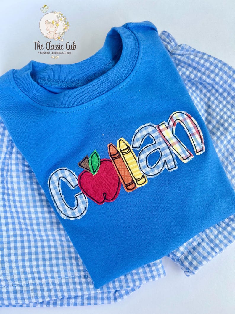 Puede incluir: Una camiseta azul para ni&ntilde;os con el nombre "Collan" bordado en un patr&oacute;n de cuadros, con una manzana y crayones. La camiseta est&aacute; superpuesta a una camisa a cuadros azul y blanca. El logotipo de The Classic Cub est&aacute; en la esquina superior izquierda.