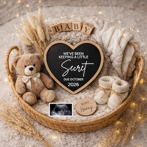 Könnte beinhalten: Ein Weidenkorb enthält einen Teddybären, eine herzförmige Tafel, Babyschuhe und Holzblöcke mit der Aufschrift "BABY". Auf der Tafel steht "WE'VE BEEN KEEPING A LITTLE SECRET DUE OCTOBER 2026". Ein Ultraschallbild und eine Holzscheibe sind ebenfalls vorhanden.