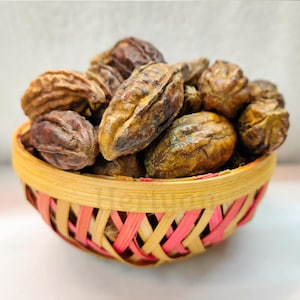 May include: A woven basket filled with brown, oblong-shaped dried fruits. The basket has a yellow and pink woven pattern. The fruits have a textured surface and varying shades of brown and yellow.