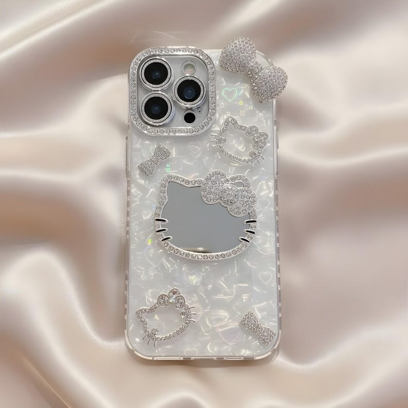 May include: A white iPhone case with a pearlized finish, adorned with sparkling rhinestones. The case features a Hello Kitty design, bows, and a camera lens frame, all embellished with crystals. The overall design is elegant and feminine.