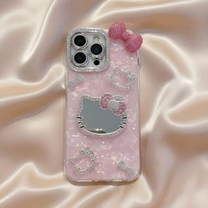 May include: Pink phone case with a Hello Kitty design. The case features a mirror, a bow, and several Hello Kitty outlines, all embellished with rhinestones. A rhinestone-studded bow is at the top. The camera lens is surrounded by rhinestones.
