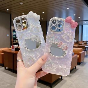 May include: Two decorative phone cases. One is white with a silver bow, the other is pink with a pink bow. Both cases feature a Hello Kitty design with sparkling embellishments around the camera and the Hello Kitty face.