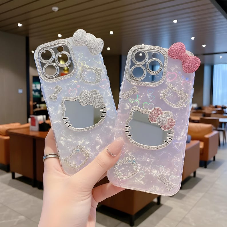 May include: Two decorative phone cases. One is white with a silver bow, the other is pink with a pink bow. Both cases feature a Hello Kitty design with sparkling embellishments around the camera and the Hello Kitty face.