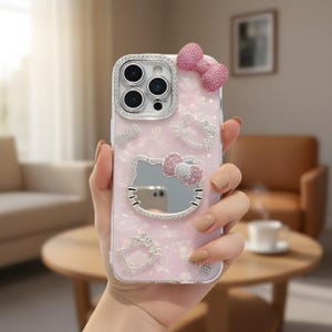 May include: Pink phone case with a Hello Kitty design. The case features a mirror, bow, and rhinestone accents. The camera lens is surrounded by a ring of rhinestones. The case has a pearlescent finish.