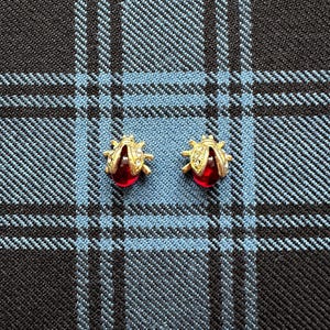 May include: Two ladybird stud earrings. Each earring has a gold-coloured body with clear crystal accents and a red, teardrop-shaped centre. The earrings are set against a blue and black plaid background.