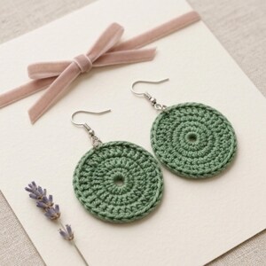 May include: A pair of handmade green crochet earrings. Each earring is a circular design with a central hole and a silver-colored hook. The earrings are displayed on a white surface with a pink ribbon and lavender sprig.