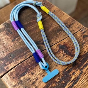 Pode incluir: Um cordão de corda azul-claro com um conector em forma de T na base. O cordão é acentuado com seções de fios enrolados em roxo, azul-escuro e amarelo, e inclui um segmento de corda estampada em azul e amarelo com um enrolamento amarelo.