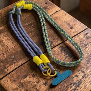 Puede incluir: Un cordón para el cuello con una cuerda larga estampada en azul y amarillo, combinada con secciones de cuerda gris sólida. El diseño incluye detalles de plástico amarillo brillante, dos anillos circulares amarillos y un accesorio azul en forma de T.