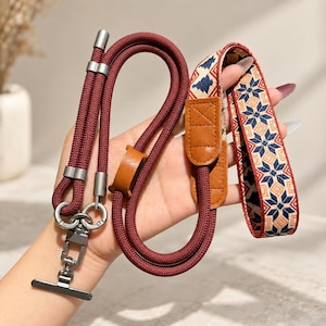 Puede incluir: Una correa para cámara con una cuerda trenzada de color rojo oscuro conectada a una sección de tela tejida plana con un patrón geométrico de estrellas azules, bronceadas y rojas. Incluye detalles de cuero marrón y herrajes de metal gris mate.