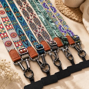May include: A collection of six fabric phone lanyards featuring various colourful geometric and floral patterns. Each strap includes a brown faux leather tab, a metal adjustable buckle, and a black metal lobster clasp attached to a black phone anchor.