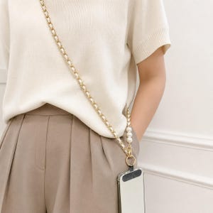 Pode incluir: Uma alça de corrente de metal dourado para telemóvel, com um design entrançado e detalhes em material branco. A alça inclui uma secção de três contas esféricas brancas perto da base, ligada a um anel circular e a uma capa de telemóvel.