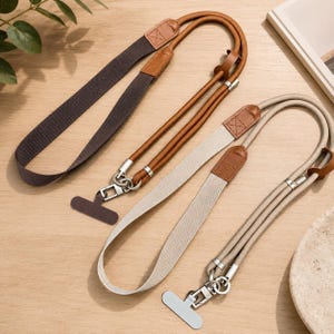 May include: Two adjustable phone lanyards featuring a combination of woven fabric straps and round cord sections. One lanyard is dark brown with tan leather accents, and the other is light beige with tan leather accents, both with metal clips.