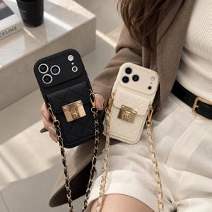 Op de afbeelding: Twee telefoonhoesjes, één zwart en één wit, met een gewatteerde textuur, een rechthoekig vakje met een goudkleurige metalen sluiting en een afneembare goudkleurige kettingriem verweven met bijpassend zwart of wit materiaal.