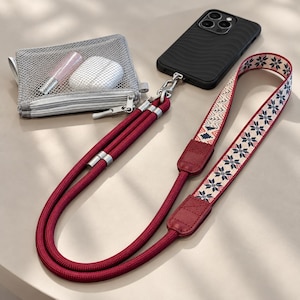 May include: A black smartphone with a wavy texture case attached to a long, dark red rope lanyard. The lanyard features a section of cream-coloured fabric with a dark blue and red snowflake pattern. A small grey mesh zippered pouch contains a white case.