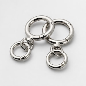 May include: Two silver-toned metal spring gate rings, each featuring a large circular loop with a spring-loaded opening mechanism connected to a smaller, secondary circular ring via a swivel joint, showcasing a polished, reflective metallic finish.
