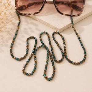 Op de afbeelding: Een lange brillenketting met afwisselend gefacetteerde blauwgroene en metallic bronzen kralen. De ketting is bevestigd aan de pootjes van een zonnebril met schildpadpatroon, waarbij de kralen in een doorlopend, herhalend patroon zijn gerangschikt.