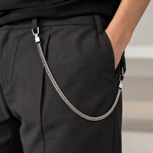 May include: A silver-toned metal chain with a herringbone pattern attached to the belt loops of black trousers. The chain features rectangular clasps at both ends, creating a curved drape across the front of the dark fabric garment.