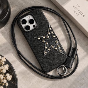 Puede incluir: Una funda de teléfono negra texturizada con un diseño de estrella grande que presenta tachuelas metálicas plateadas. La funda incluye una correa larga de cordón negro con anillos metálicos plateados en la base para llevar el dispositivo.