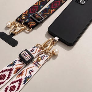 May include: Two adjustable phone straps with woven geometric patterns in black, white, red, and blue. Each strap features gold-tone metal lobster clasps, a white faux pearl accent, and a black plastic slider. One strap is attached to a black smartphone.