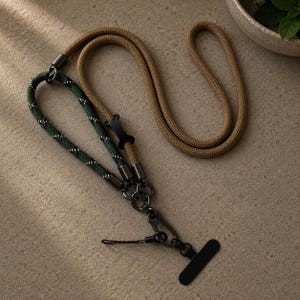 Peut inclure: Une lanière bicolore composée d'une section en corde beige unie et d'une section en corde vert foncé à motifs pointillés blancs. Les deux cordes sont reliées par des pièces métalliques sombres, incluant un clip central, un mousqueton et un embout noir.