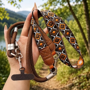 May include: A brown rope lanyard featuring a flat, woven fabric strap with a geometric pattern in orange, black, and white. The strap includes brown faux leather accents and silver-toned metal swivel hooks, with a dark brown fabric phone anchor tab.