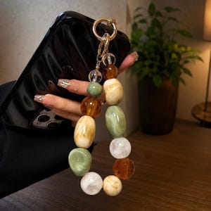 Op de afbeelding: Een telefoonbedel met een verzameling gladde, veelkleurige kralen in tinten amber, saliegroen, crème en helder wit. De kralen zijn in een decoratief patroon aan elkaar geregen en bevestigd aan een goudkleurige metalen ringsluiting.
