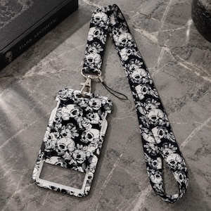 May include: A black and white lanyard with a repeating skull and floral pattern attached to a matching vertical ID badge holder. The badge holder features a clear rectangular window for displaying identification cards and a metal swivel hook.