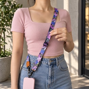 Peut inclure: Une bandoulière pour téléphone avec un motif de feuilles tropicales aux tons violet, bleu et noir. La sangle comprend une boucle de réglage en plastique noir, une patte en cuir marron et des mousquetons pivotants noirs fixés à une coque.