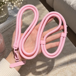 May include: A light pink phone lanyard featuring a braided cord design with matching pink plastic connectors and a rectangular attachment tab. The accessory includes a swivel clip mechanism for securing a mobile device, presented in a solid pastel colour.