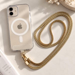 May include: A clear smartphone case with a white circular MagSafe magnetic ring and an Apple logo visible on the back. The case is attached to a long, thick, gold-toned metal chain strap with a polished finish, featuring a rectangular connector.