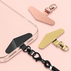 May include: Three adhesive phone tether tabs with metal swivel clips, including one black tab attached to a clear phone case with a black lanyard, one rose gold tab, and one gold tab, all featuring a triangular shape for secure device attachment.