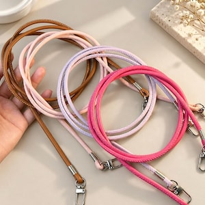 Puede incluir: Un conjunto de cuatro correas redondas tipo cordón para bolso en colores marrón claro, rosa pálido, lavanda y rosa intenso. Cada correa cuenta con cierres de mosquetón de metal plateado en ambos extremos, asegurados con casquillos metálicos a juego.