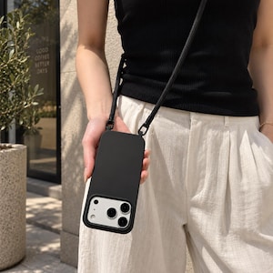 Op de afbeelding: Een effen zwarte smartphonehoes met een bijpassende zwarte crossbody-riem bevestigd aan de bovenhoeken. De hoes heeft een rechthoekige uitsparing voor de cameramodule, waardoor de zilverkleurige lenzen en flitser van de telefoon zichtbaar zijn.